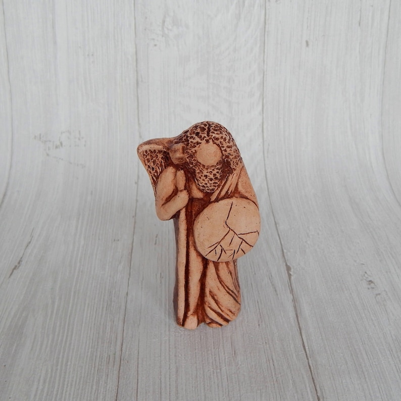 May include: A terracotta figurine of a person holding a shield and a staff. The figurine is brown and has a detailed design.