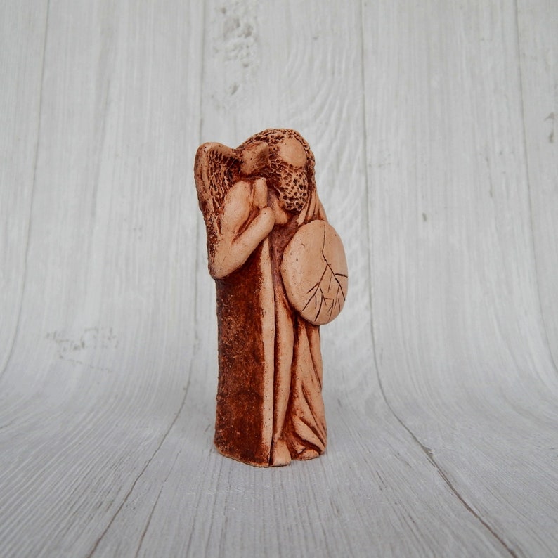 May include: A ceramic figurine of an angel holding a shield with a tree design. The angel is brown and has a textured surface.