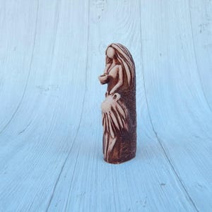 May include: A ceramic figurine of a woman with long hair, wearing a flowing dress and holding a small object in her hand. The figurine is painted in shades of brown and beige.