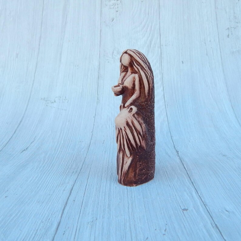 May include: A ceramic figurine of a woman with long hair, wearing a flowing dress and holding a small object in her hand. The figurine is painted in shades of brown and beige.