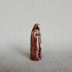 May include: A brown ceramic figurine of a person wearing a long robe and holding a harp. The figurine is standing on a white surface.