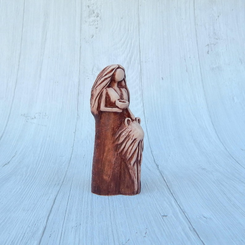 May include: A ceramic figurine of a woman holding a small bowl in her hands. She is wearing a long, flowing dress and has long, flowing hair. The figurine is painted in shades of brown and white.