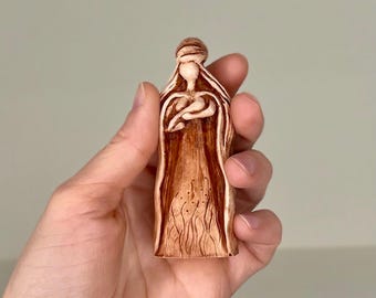 Könnte beinhalten: Eine kleine Keramikfigur einer Frau, die ein Baby in ihren Armen hält. Die Figur ist in Braun- und Weißtönen bemalt.