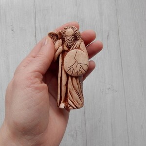 May include: A hand-painted ceramic figurine of a figure holding a shield with a tree design. The figurine is made of a light brown clay and has a rustic, earthy look.