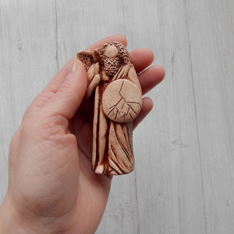 May include: A hand-painted ceramic figurine of a figure holding a shield with a tree design. The figurine is made of a light brown clay and has a rustic, earthy look.