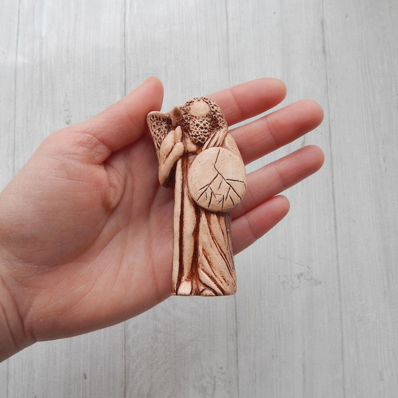 May include: A small, hand-crafted ceramic figurine of an angel holding a shield with a tree design. The angel is wearing a long robe and has wings. The figurine is painted in shades of brown.