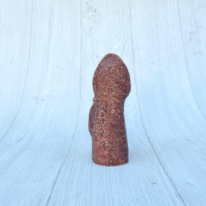May include: A brown ceramic sculpture of a person standing with their arms at their sides. The sculpture has a rough, textured surface.