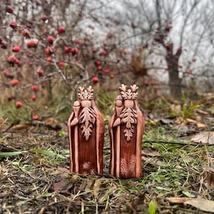 Puede incluir: Dos pequeñas figuritas de cerámica pintadas a mano que representan a personas con coronas y túnicas de hojas, de pie sobre un lecho de hojas marrones y hierba verde. Las figuritas son de color marrón rojizo.