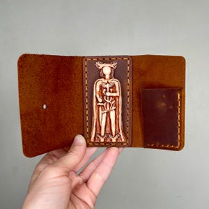 May include: A brown leather wallet with a carved image of a figure holding a staff and a snake. The wallet has a flap that closes with a snap.