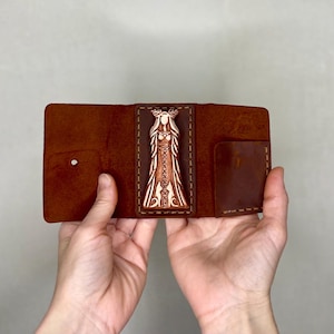 May include: A brown leather wallet with a carved wooden panel featuring a woman with a crown of antlers. The wallet is open and the panel is visible.