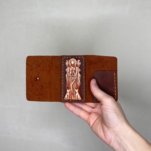 May include: A brown leather wallet with a carved design of a woman with two cats on the front. The wallet is closed and being held in a hand.