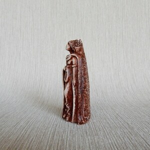 May include: A brown ceramic figurine of a woman wearing a long robe and a crown. The figurine is standing on a light brown surface.