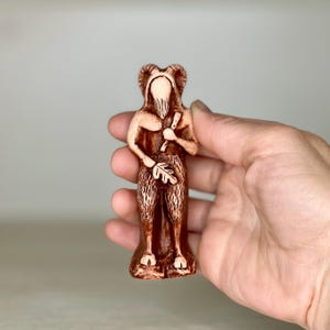 May include: A small, brown ceramic figurine of a goat-like creature with horns, a beard, and a leaf in its hand. The figurine is standing on a base and has a detailed design.