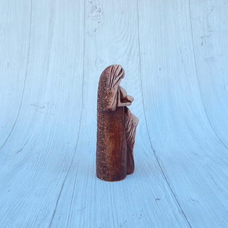 May include: A ceramic sculpture of a woman holding a child. The sculpture is painted in shades of brown and has a rustic, earthy feel.
