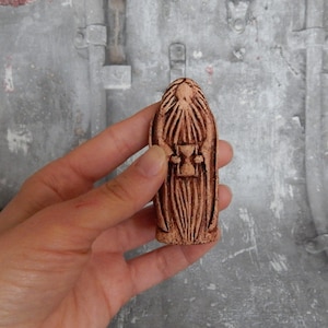 May include: A small, brown ceramic figurine of a person with a long beard and a robe. The figurine is holding a small hourglass in their hands.