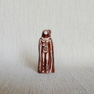 May include: A brown ceramic figurine of a person wearing a long robe and holding a lyre. The figurine has a white face and is standing on a white background.