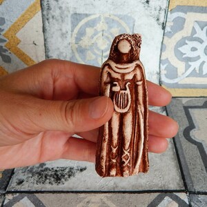 May include: A small, brown and white ceramic figurine of a person holding a lyre. The figurine is wearing a long robe and has a detailed face.