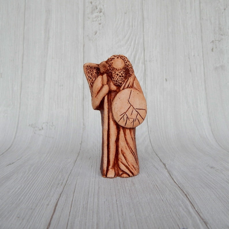 May include: A terracotta figurine of a person holding a shield and a staff. The figurine is standing on a white wooden surface.
