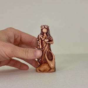 May include: A small, hand-painted ceramic figurine of a woman with a crown of leaves, holding a cup. The figurine is brown and has a detailed design.