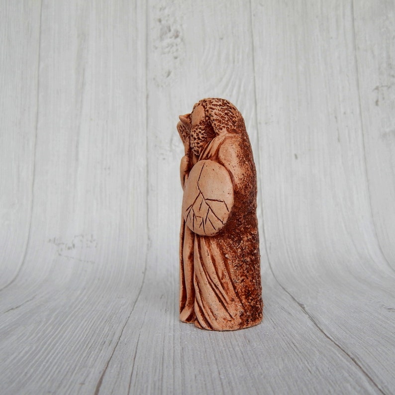 May include: A terracotta figurine of a person holding a shield with a tree design. The figurine is standing on a white wooden surface.