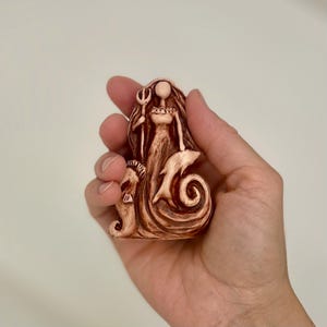 Könnte beinhalten: Braune Figur einer Meerjungfrau mit einem Dreizack, mit einem Seepferdchen und einem Delfin. Die Meerjungfrau hat lange Haare und trägt ein Kleid. Die Figur wird in einer Hand gehalten.