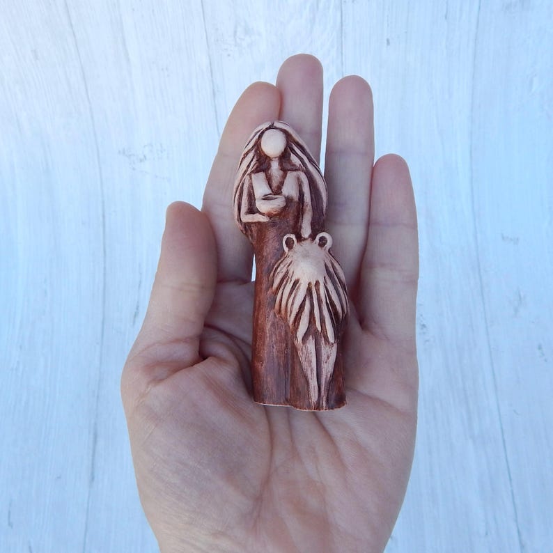 May include: A hand-painted ceramic figurine of a woman holding a smaller figurine of a child. The woman is wearing a long dress and has long hair. The child is wearing a dress and has short hair. The figurines are painted in shades of brown and beige.