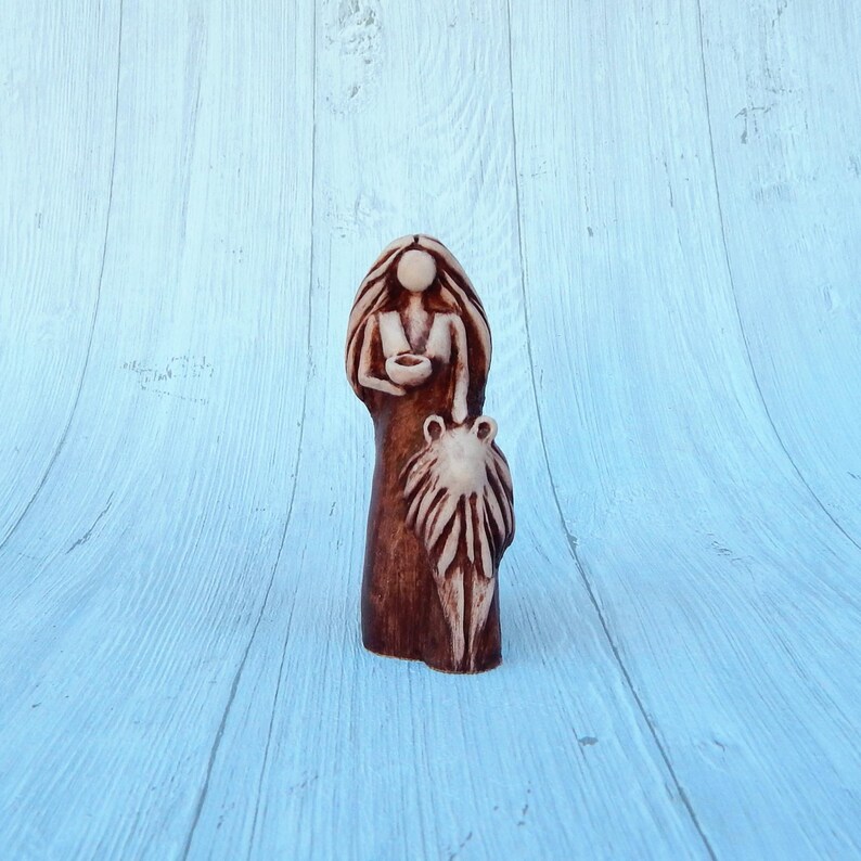 May include: A ceramic figurine of a woman holding a bowl in her hands. She is wearing a long dress and has long hair. The figurine is painted in shades of brown and white.