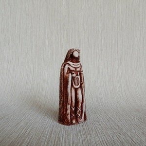 May include: A brown ceramic figurine of a person wearing a long robe and holding a lyre. The figurine is standing on a light brown surface.