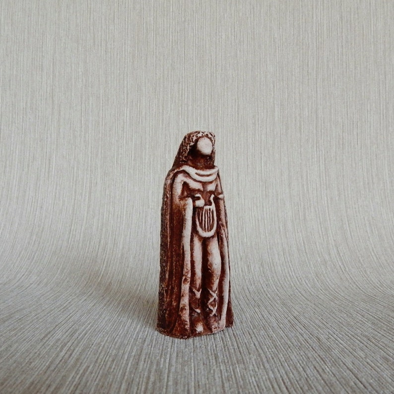 May include: A brown ceramic figurine of a person wearing a long robe and holding a lyre. The figurine is standing on a light brown surface.