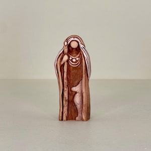 Könnte beinhalten: Kleine, braune Figur einer bekleideten Figur mit Stab. Die Figur hat eine weiße Kopfbedeckung und eine Halskette. Die Figur steht aufrecht und scheint aus einem geschnitzten Material zu bestehen.