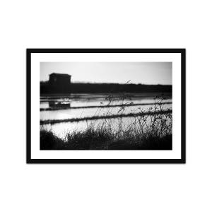 Peut inclure: Photographie en noir et blanc d'un marais salant avec un petit bâtiment au loin. L'image est prise d'un angle bas, avec une mise au point sur les hautes herbes au premier plan.