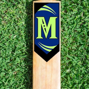 May include: A cricket bat made of wood, with a light-coloured handle and a rectangular face. The face has a blue and black design with a large, lime green letter "M" and curved green accents. The bat is placed on green grass.