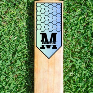 May include: A cricket bat made of wood, with a light brown colour. It has a black and blue hexagonal design and the name "RICHARDSON" below a large black "M". The bat is on green grass.