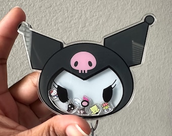 May include: A black and white character with a pink nose and skull on its head. The character is surrounded by small colorful characters. The character is attached to a clear plastic badge holder with a metal clip.