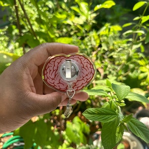 May include: A clear heart-shaped badge holder with a red and white brain design. The holder has a metal clip and a retractable cord.