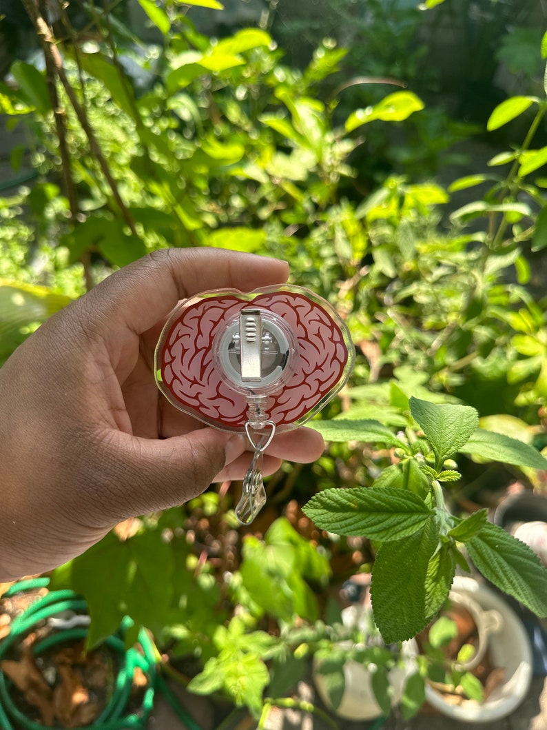 May include: A clear heart-shaped badge holder with a red and white brain design. The holder has a metal clip and a retractable cord.