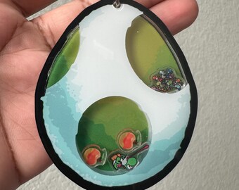 May include: A green and white egg-shaped keychain with a clear plastic insert containing a small green Yoshi figure and red and orange coins. The keychain has a silver chain attached to the top.