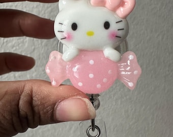 May include: A white and pink Hello Kitty badge reel. The character has a pink bow, rosy cheeks, and black whiskers. The reel is attached to a pink candy-shaped charm with white polka dots. The reel has a clear plastic strap and a metal clip.