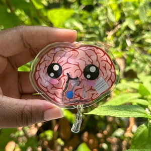 May include: A clear plastic keychain shaped like a brain with a cute cartoon face. The brain is pink and has a bandage on it. The keychain has a metal ring for attaching to keys.