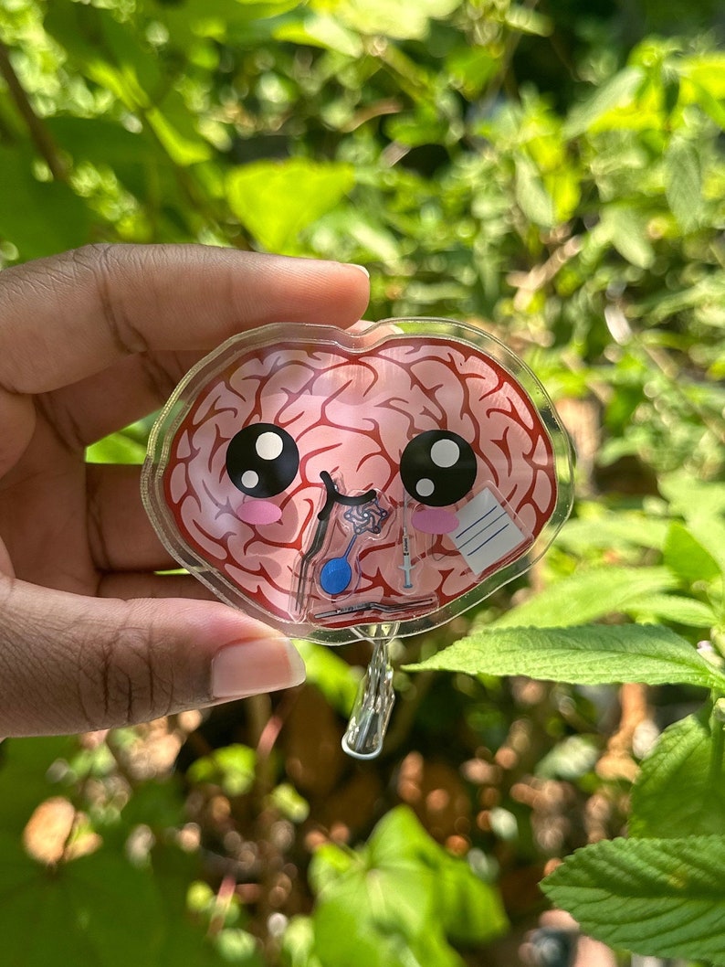 May include: A clear plastic keychain shaped like a brain with a cute cartoon face. The brain is pink and has a bandage on it. The keychain has a metal ring for attaching to keys.