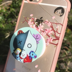 May include: A pink iPhone case with a cherry blossom design and a pop socket featuring a black and white character with other characters surrounding it. The pop socket is green and white.