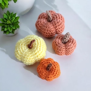 May include: Four small, handmade crochet pumpkins in shades of dusty rose, pale yellow, and bright orange. Each pumpkin features a textured, ribbed surface and a short, brown crocheted stem attached to the top centre of the rounded body.