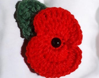 May include: A handcrafted crochet poppy flower brooch featuring vibrant red yarn petals and a single dark green leaf. The centre of the flower is accented with a small, smooth, round black bead, providing a distinct contrast to the textured red yarn.