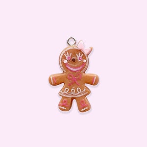 Può includere: Un ciondolo a forma di omino di pan di zenzero con dettagli in glassa rosa e un fiocco rosa. Il ciondolo è in ceramica e ha un foro nella parte superiore per appenderlo.
