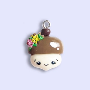 May include: A brown and white acorn charm with a smiling face and a flower crown. The charm has a silver loop for hanging.