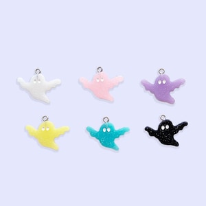 May include: A set of six small ghost-shaped charms, each featuring a glittery finish and a metal jump ring at the top. The ghosts are colored white, light pink, light purple, yellow, light blue, and black, all with two small circular eye cutouts.