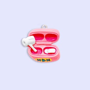 May include: A pink charm shaped like an open charging case for wireless earbuds. The interior is bright pink, containing two white earbud replicas. The front of the case features a yellow horizontal bar with small blue and green playback icons.