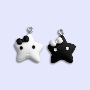 May include: Two star-shaped charms with a smiling face and a bow. One charm is white with a black bow and the other is black with a white bow.