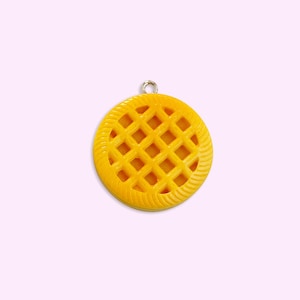 Peut inclure: Un charm jaune et circulaire ressemblant à une tarte, avec un dessus en treillis et un bord texturé. Le charm a une boucle de couleur argentée en haut pour l'attacher à un collier ou un bracelet. Le fond est rose clair.