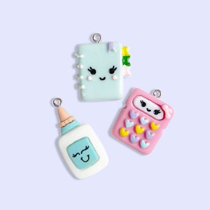 Puede incluir: Tres encantos coloridos con caras lindas. Un encanto de cuaderno azul claro, un encanto de calculadora rosa y un encanto de botella de pegamento azul claro.